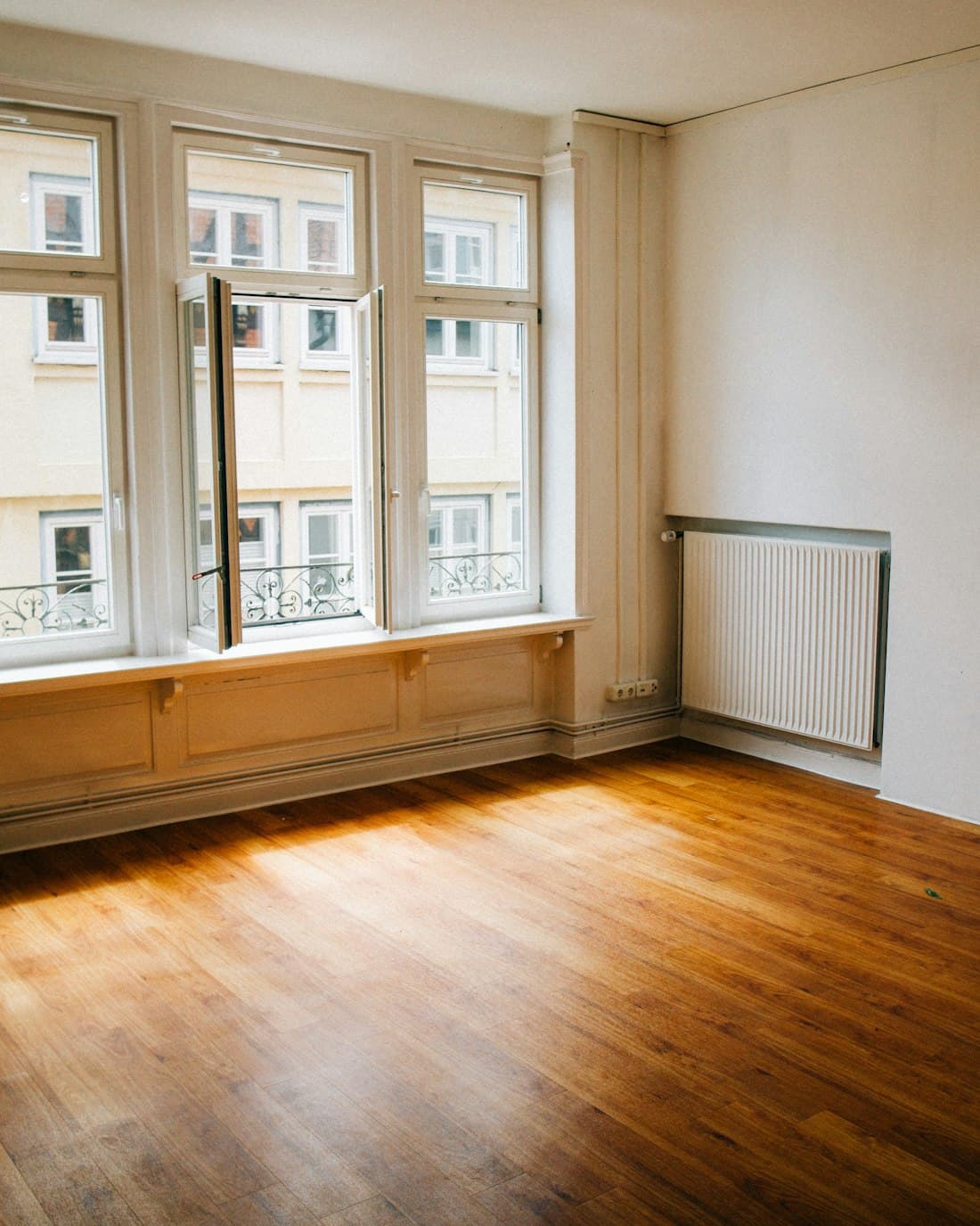 An empty apartment room with a wooden floor, tall sash windows and warm afternoon light.