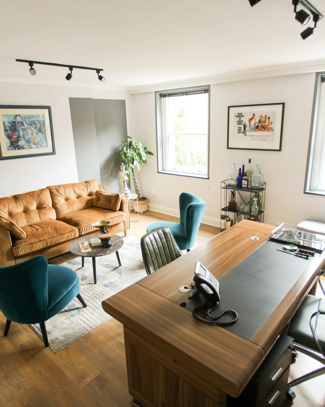 A small office with a wooden desk, leather sofa and daylight through two sash windows.
