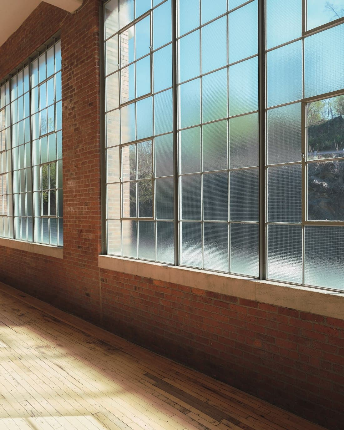 A bare room with brick walls, tall factory windows and daylight across a stripped timber floor.